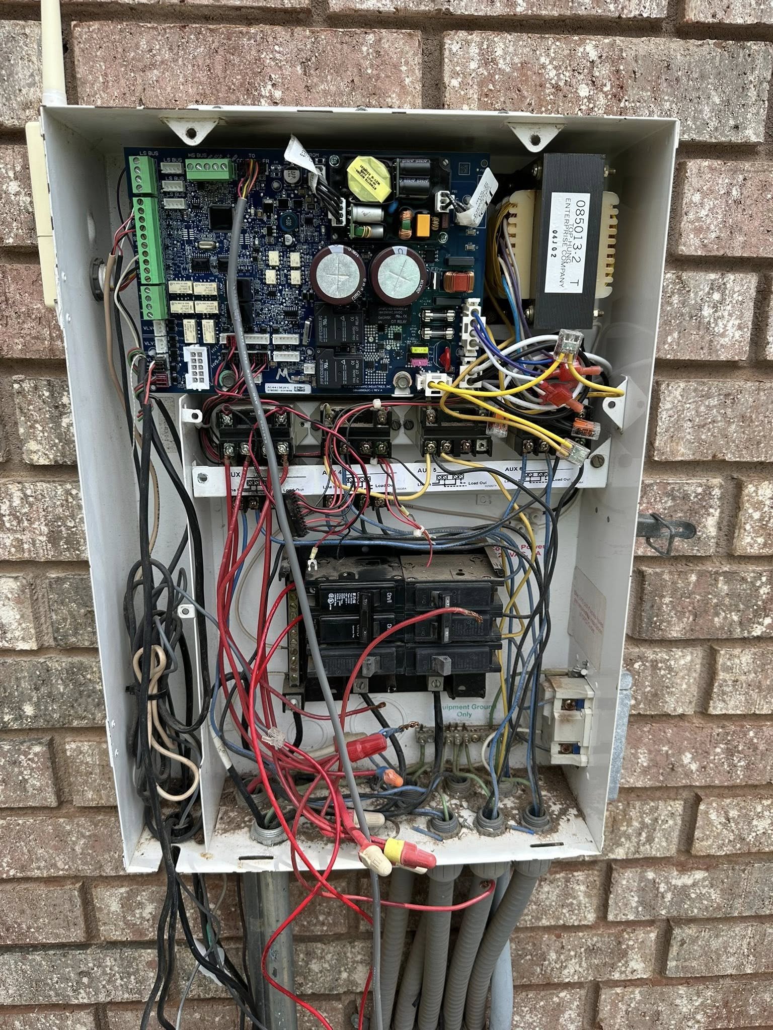 Open electrical control box containing circuit boards, wiring, relays, and electrical components mounted against a brick wall.