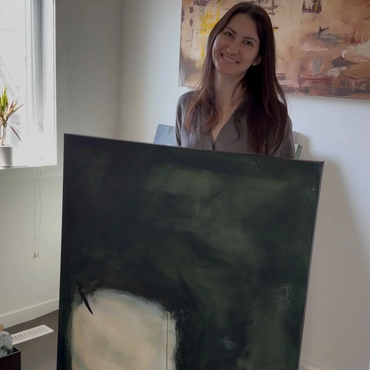 A woman with long brown hair smiling while standing behind a large abstract painting in a well-lit room with a window and a plant.