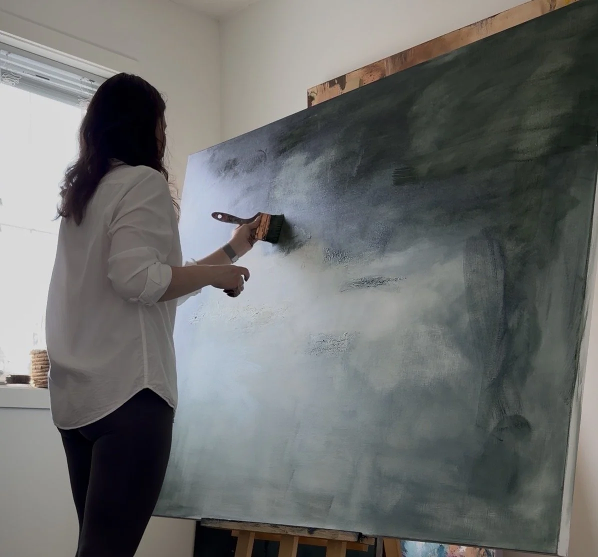Woman painting an abstract canvas with dark green and gray hues in a bright room.