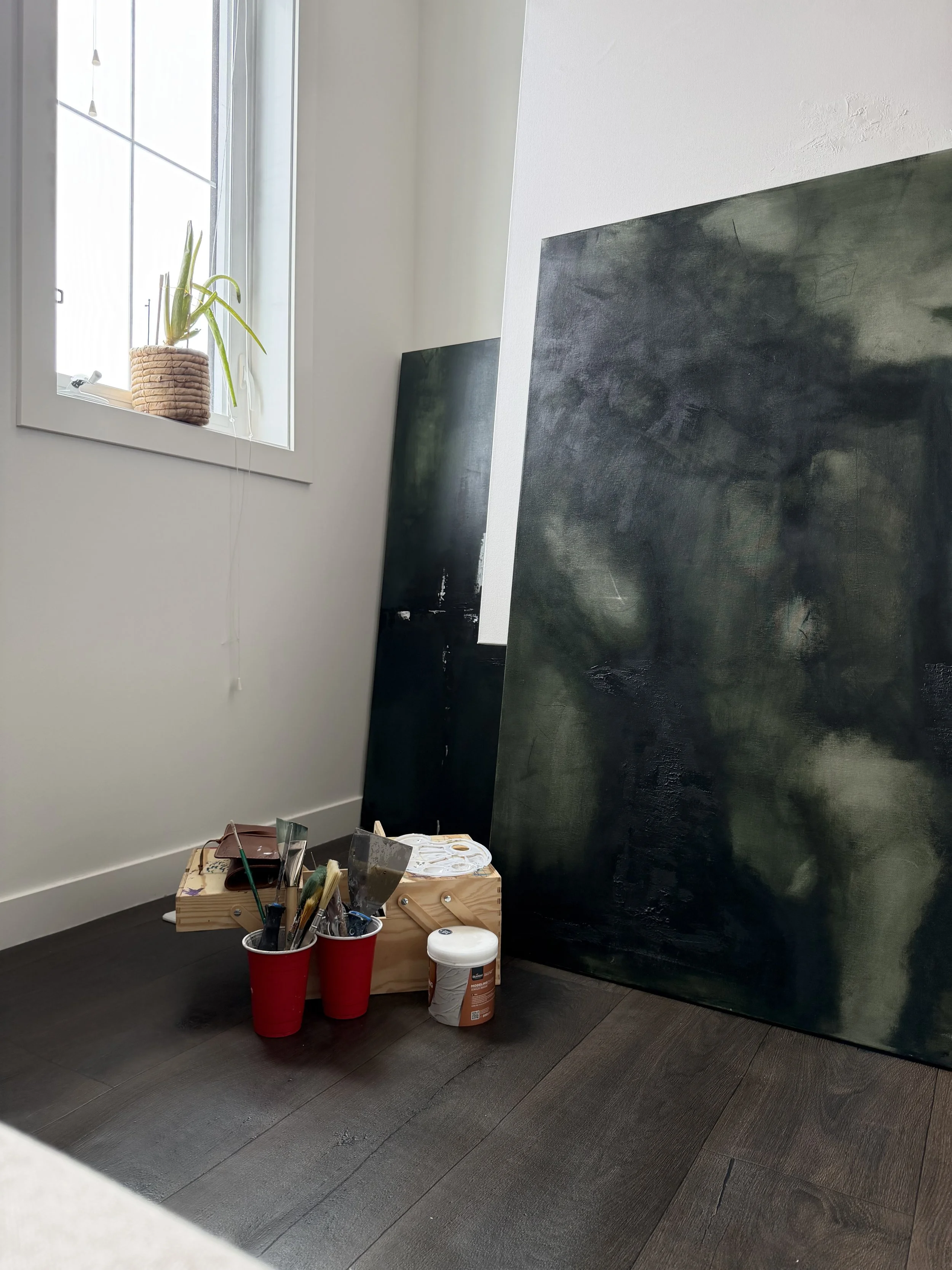 An art studio or workspace with large paintings, on a dark hardwood floor next to a window with a plant in a woven basket. Art supplies including brushes, a palette, and paint containers are on a wooden box and nearby surfaces.