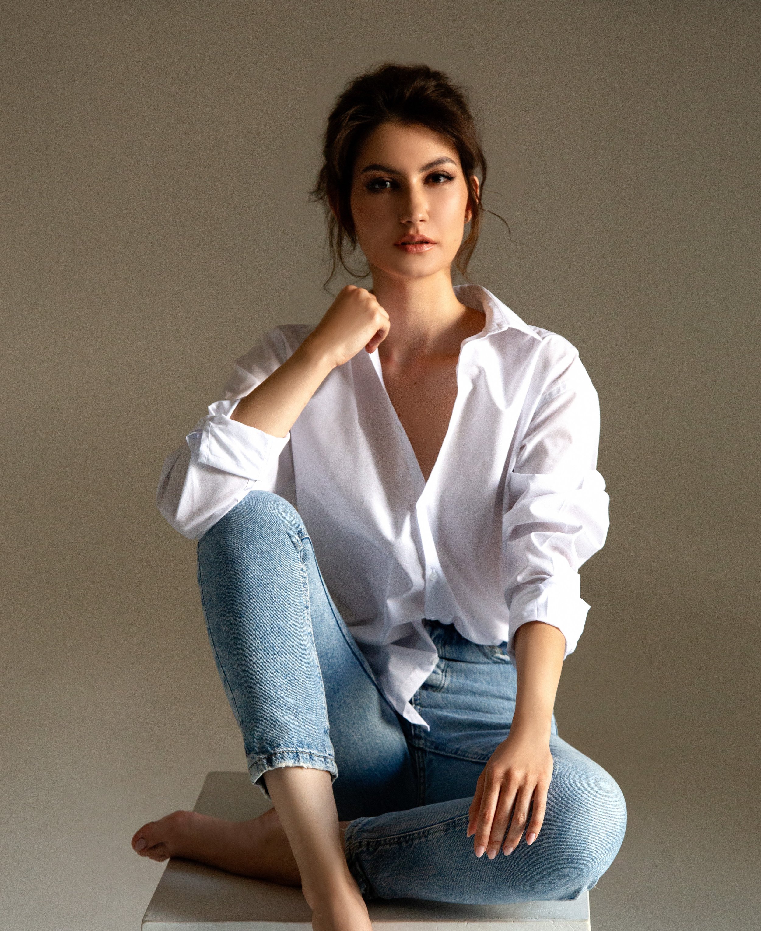 A woman with dark brown hair tied back, wearing a white button-up shirt and light blue jeans, sitting barefoot on a grey chair against a plain grey background, with her left leg crossed over her right knee and her right hand resting on her ankle.