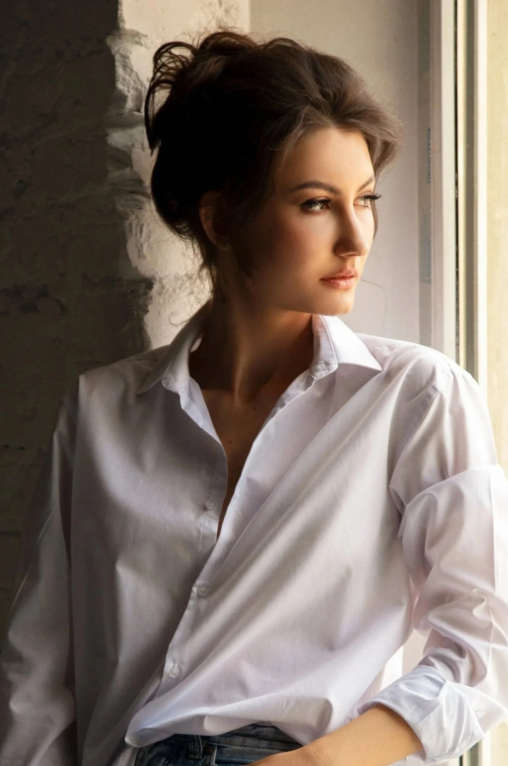 A woman with short brown hair looking out a window wearing a white shirt.