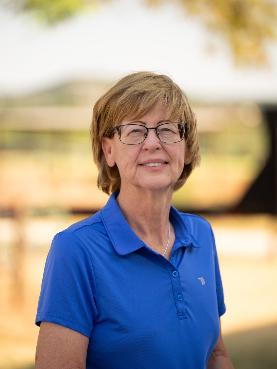 A middle-aged woman with short light brown hair and glasses, wearing a blue polo shirt, standing outdoors with blurred trees and a fence in the background.