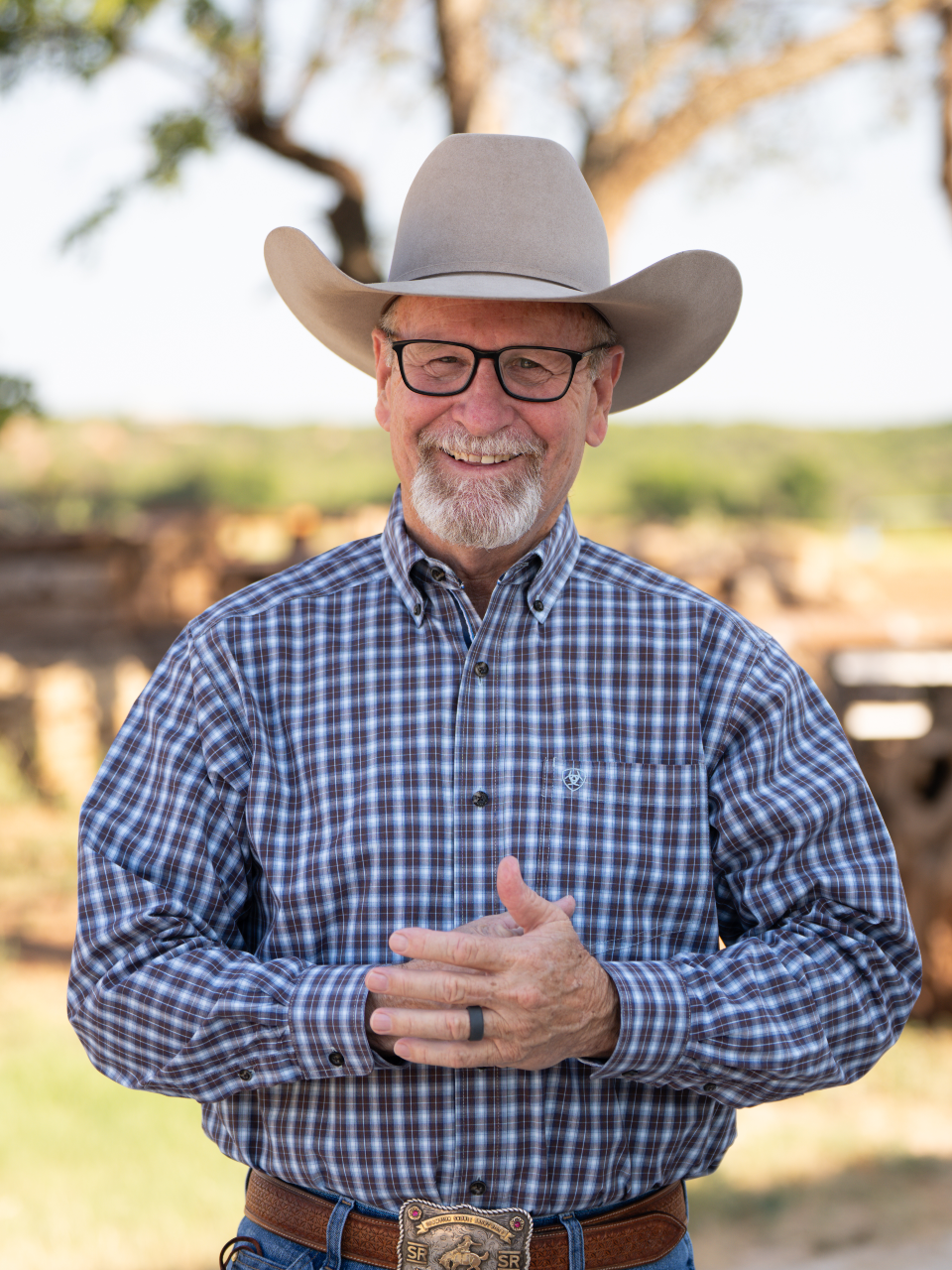 A smiling man with glasses and a white beard wearing a cowboy hat and plaid shirt standing outdoors with a tree and blurred landscape in the background.