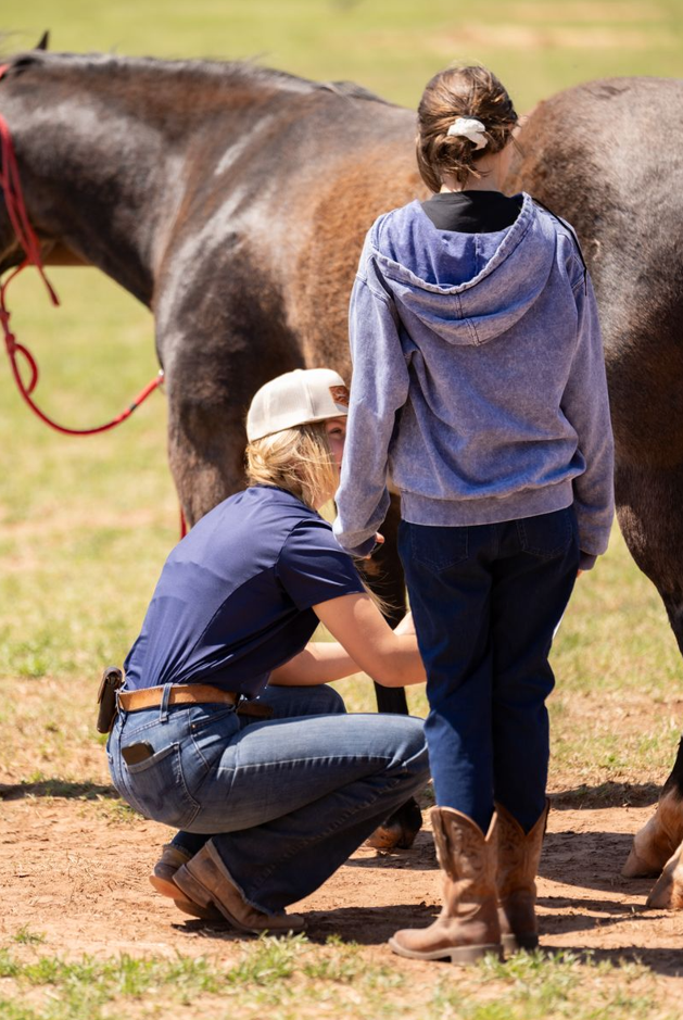 A person kneeling and a person standing next to a horse outdoors, with the person kneeling tending to the horse's leg. The person standing is wearing a gray hoodie and cowboy boots, and the person kneeling is wearing a white cap and a blue shirt.