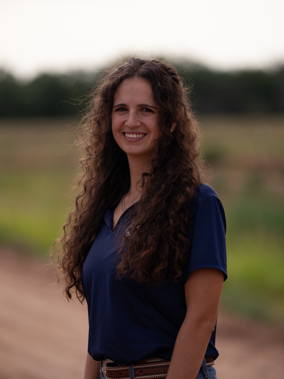 A woman with curly hair smiling and sitting outdoors in fall foliage, wearing an orange long-sleeve shirt, a white knit shawl, and blue jeans.