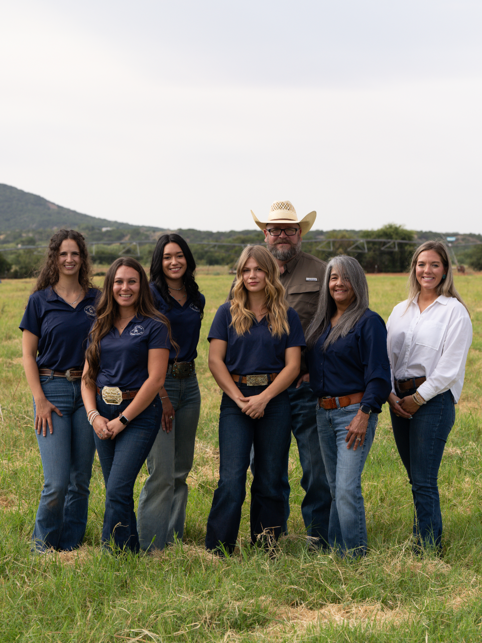 Group of six people standing outdoors on a grassy field, wearing Western-style clothing, with mountains and a cloudy sky in the background.