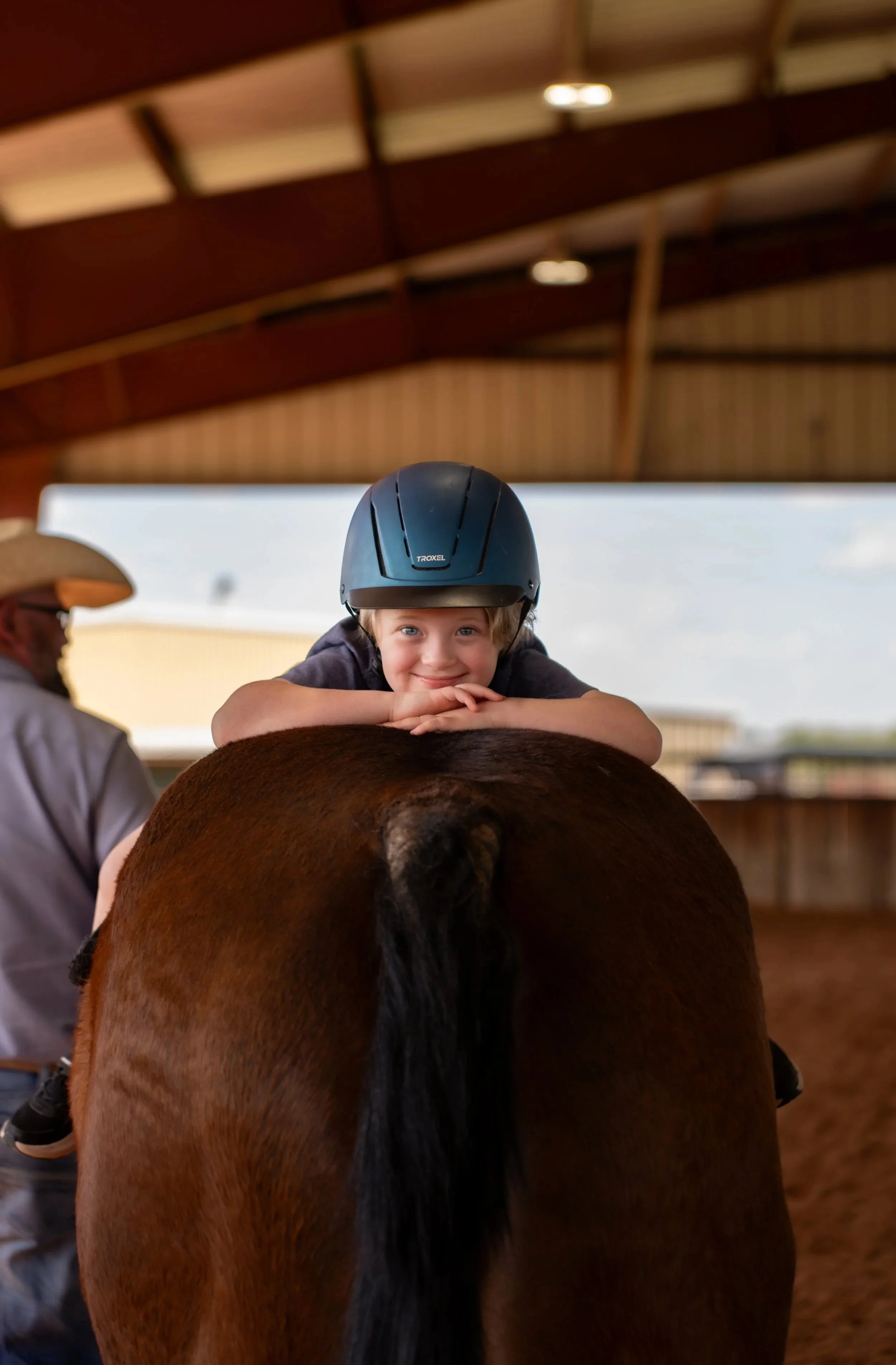 Child wearing a helmet riding a horse, with another person partly visible next to them, in an indoor riding arena.