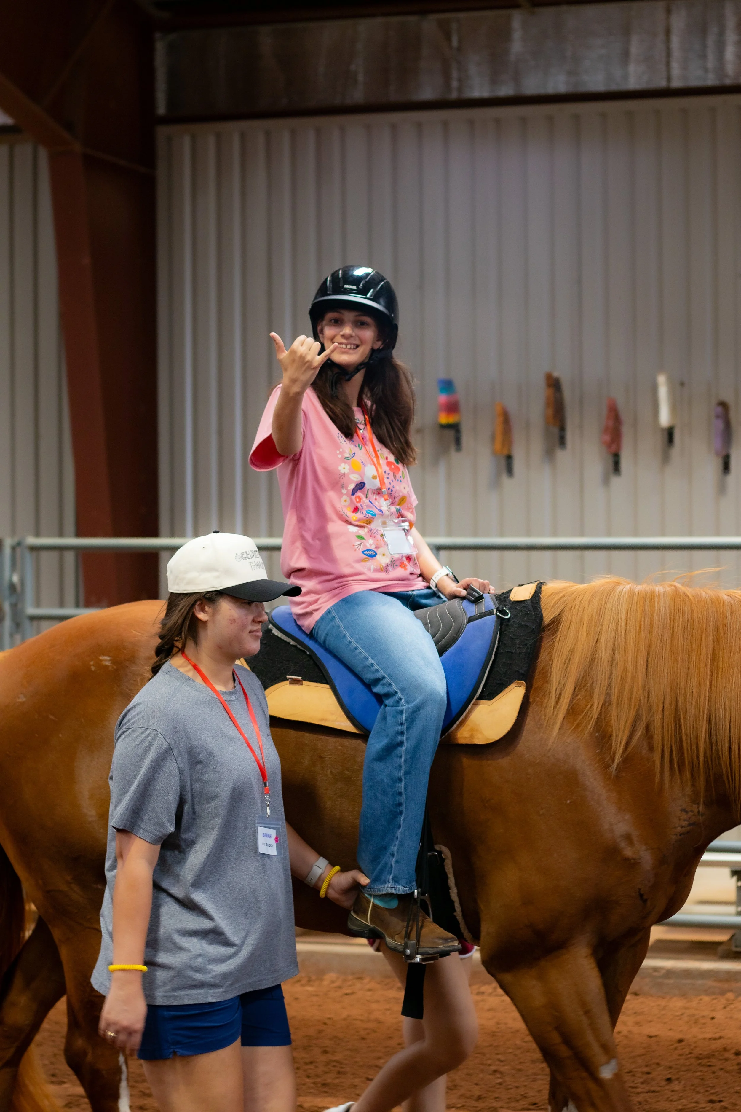 A young woman wearing a pink t-shirt, black helmet, and jeans, is riding a horse inside an indoor riding arena. She is making a shaka sign with her right hand and smiling. Two other women are walking nearby, one in a gray shirt with a name tag and a cap, and another partly visible at the horse's tail.