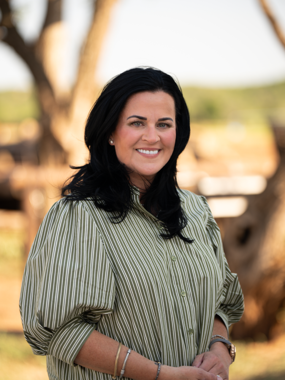 A smiling woman with black hair, wearing a green and white striped button-up shirt, standing outdoors with trees in the background.