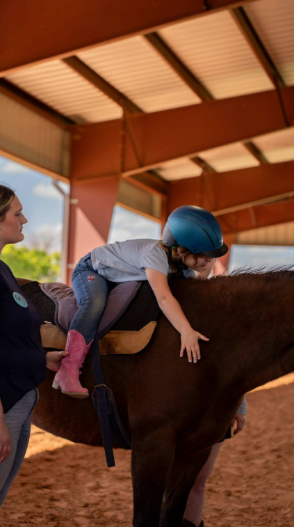 Child wearing a helmet and pink boots being guided to ride a horse at an indoor riding arena.