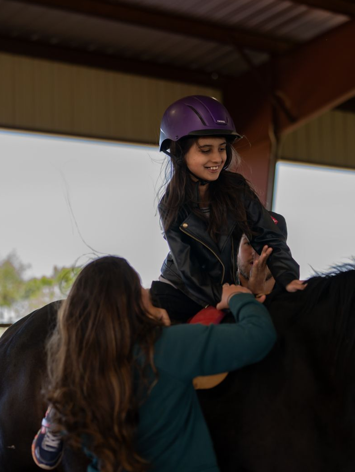 A young girl wearing a helmet and a leather jacket sits on a horse with the help of two adults inside a barn or covered riding area.