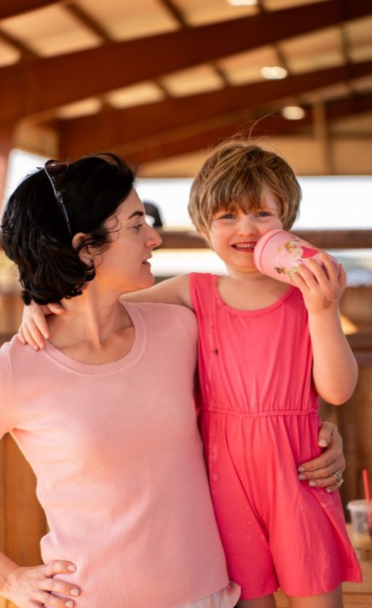 A woman and a young girl are sharing a joyful moment outdoors under a wooden pavilion. The woman has dark, wavy hair and is wearing a light pink shirt. The girl is wearing a bright pink dress and is drinking from a pink cup with a straw, smiling happily.