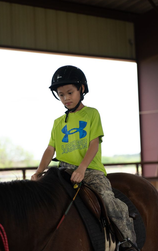 A young boy wearing a green Under Armour T-shirt and camouflage pants riding a horse indoors, with a black helmet on his head.