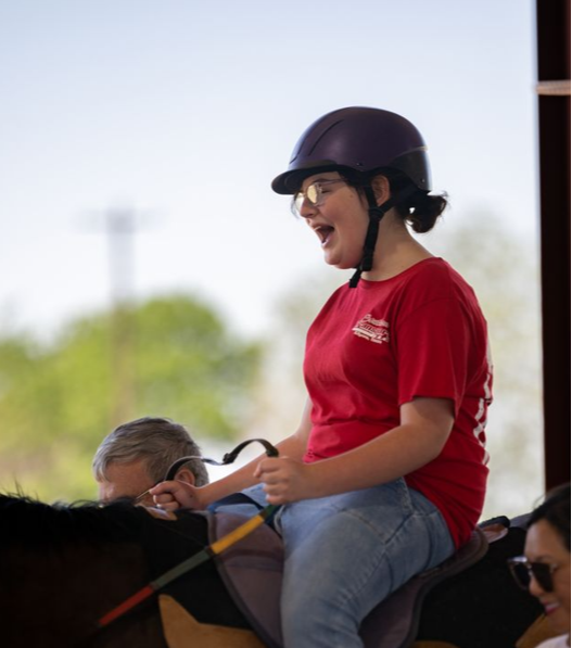 A young girl wearing a riding helmet and red shirt, smiling while riding a horse.