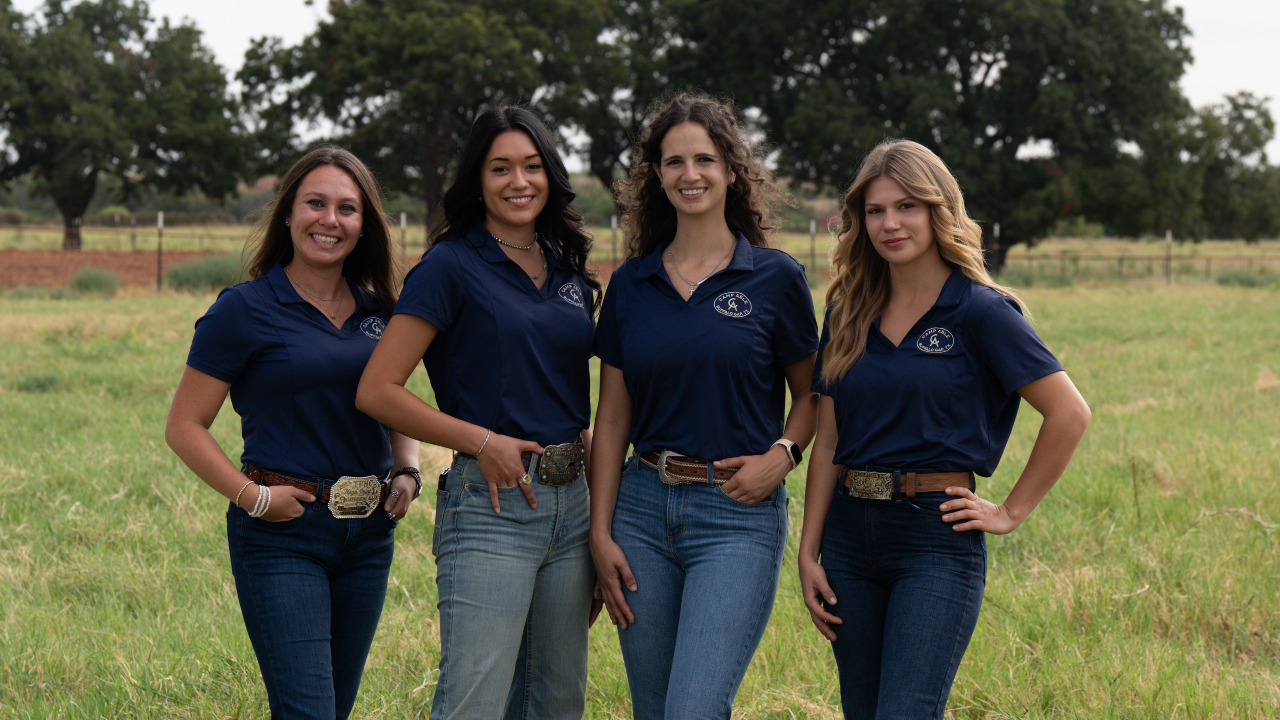Four women standing in a field, wearing matching navy blue collared shirts and jeans, smiling at the camera.
