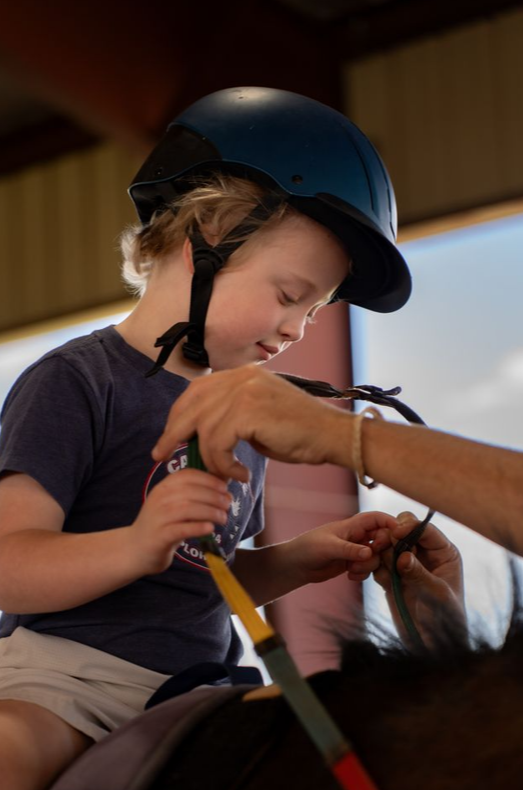Young boy wearing a blue riding helmet, sitting on a horse, receiving a helmet fitting from an adult.