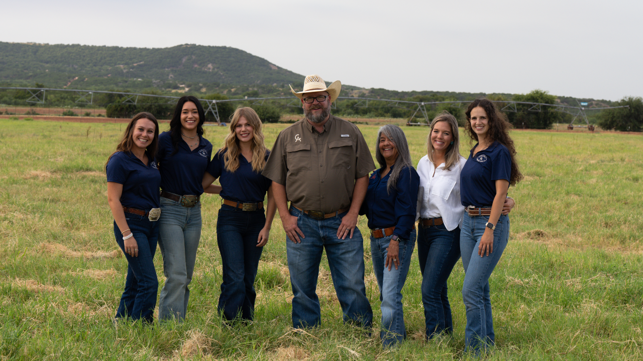 Group of six people standing outdoors on grass field with hills in background, wearing casual clothing, smiling at camera.