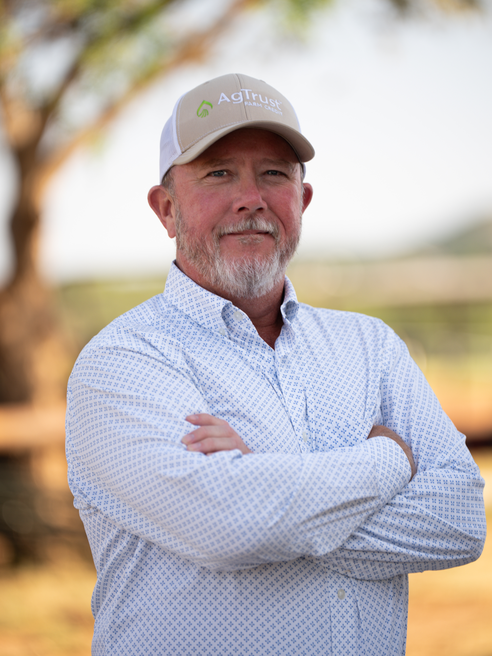 A middle-aged man with a beard and mustache wearing a beige and white baseball cap with 'AgTrust' logo, a light blue patterned button-up shirt, standing outdoors with arms crossed and a blurred natural background.