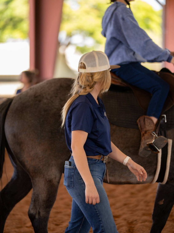 A young girl with blonde hair wearing a beige baseball cap, navy polo shirt, and jeans walking alongside a horse with a rider on its back at an indoor riding facility.