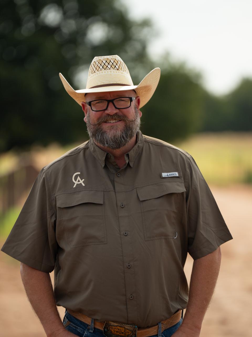 A man with a beard wearing sunglasses, a straw cowboy hat, a light blue shirt, and jeans standing next to a brown and white horse inside a barn.