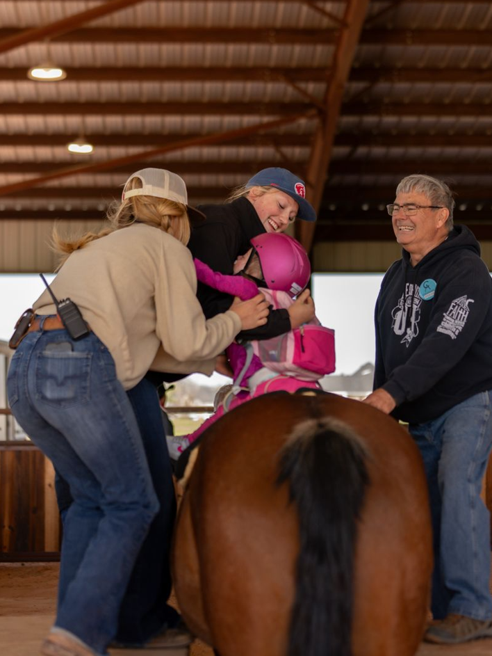 A young girl wearing a helmet rides a horse, assisted by two women and an older man at an indoor equestrian facility.