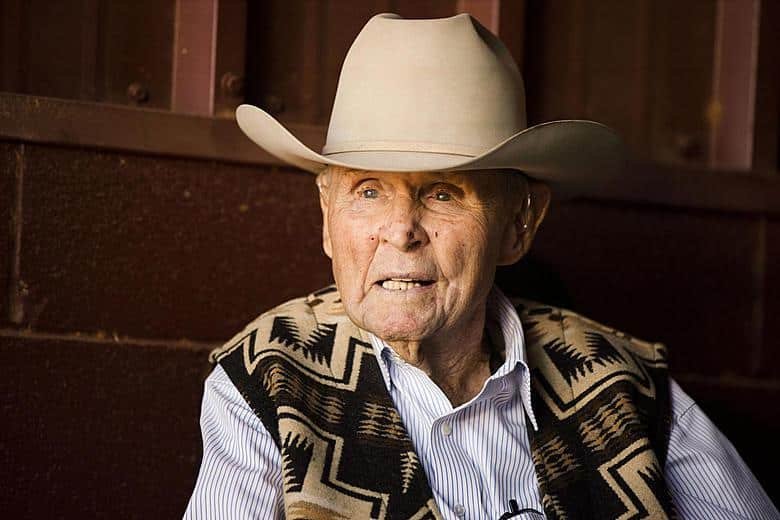 An elderly man with a cowboy hat, wearing a patterned southwestern vest over a striped shirt, sitting against a wooden background.
