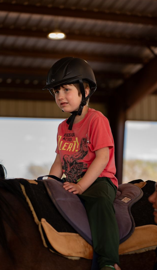 Young boy in a red t-shirt and black helmet riding a horse in an indoor riding arena.