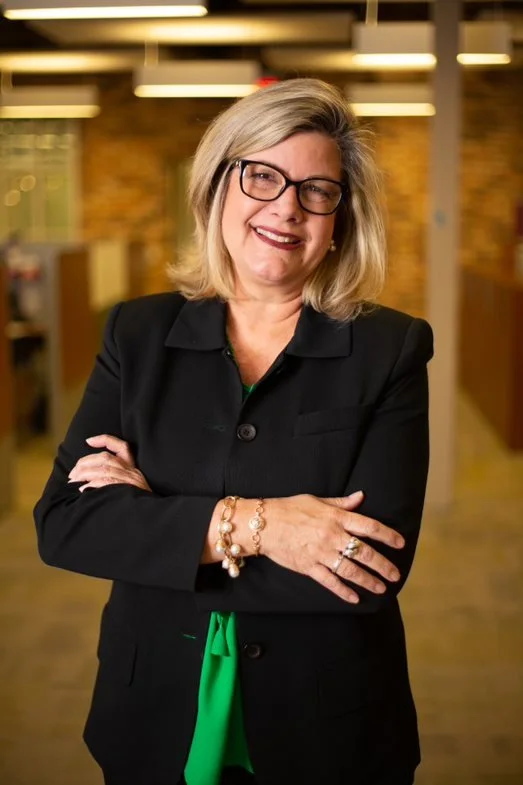 A woman with short blonde hair and glasses smiling, standing with arms crossed in a modern office setting with warm lighting.