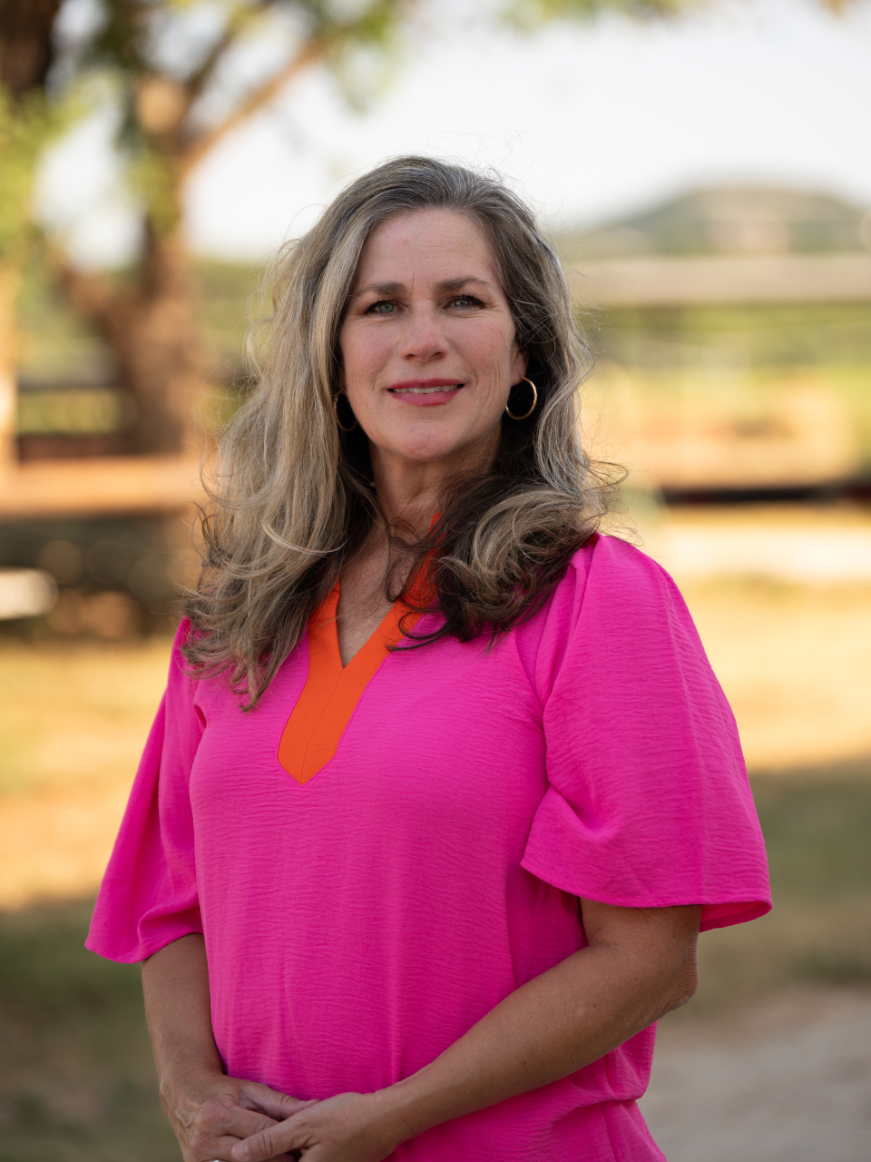 A woman with long wavy gray hair wearing a bright pink top with orange accents, standing outdoors with a blurred background of trees and a fence.