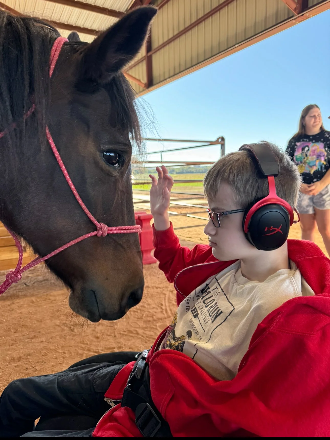A young boy wearing glasses and red headphones, wearing a red jacket and beige shirt, is sitting in a barn or stable. He is reaching out to touch the face of a dark brown horse with a pink halter. In the background, a woman and a girl are standing near the barn.