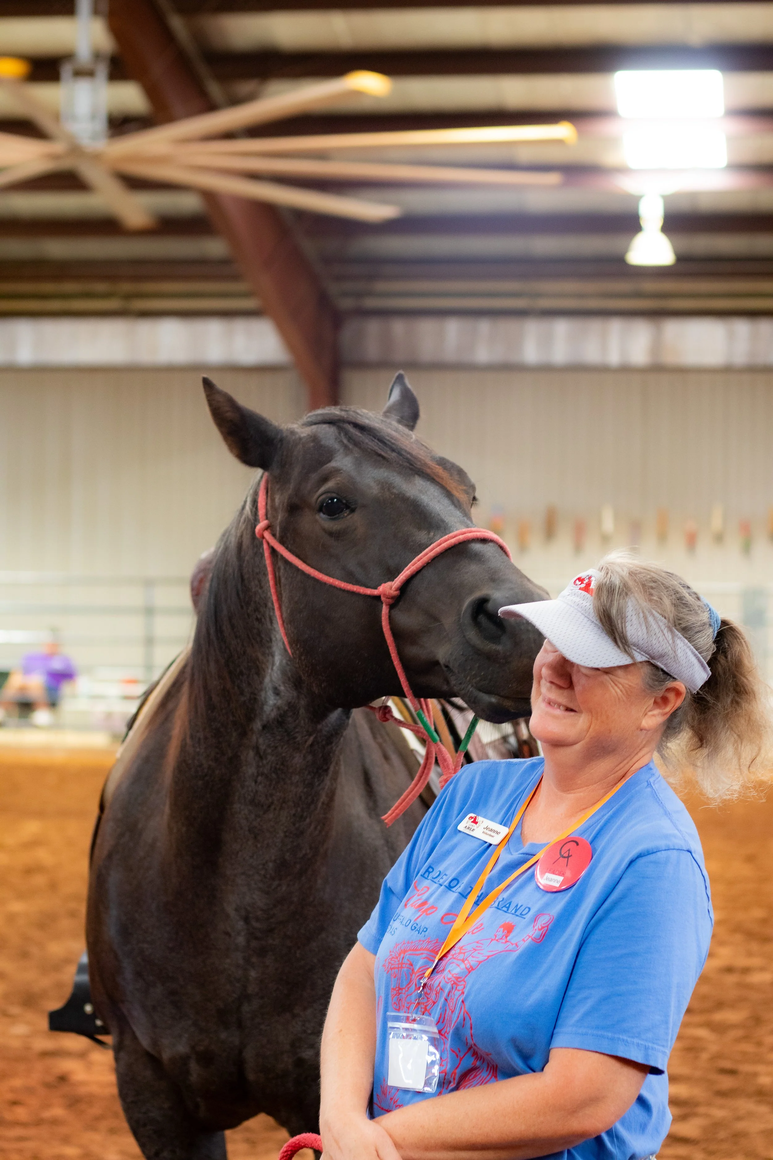 A woman smiling and wearing a blue T-shirt and a name tag, standing next to a dark horse in an indoor riding arena.