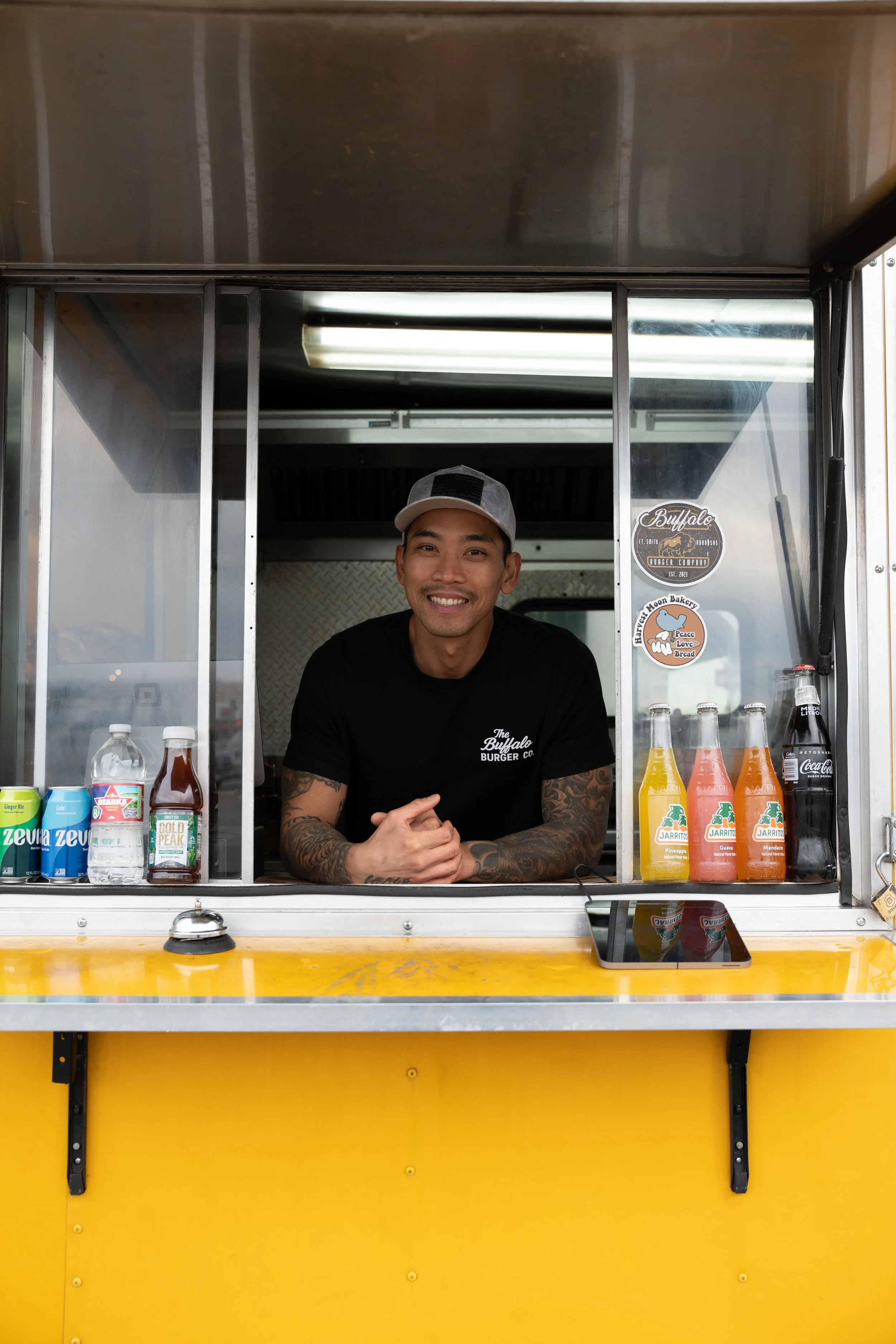 Smiling man working at a food truck, wearing a black t-shirt and a gray cap. The food truck has a yellow exterior, with bottles of drinks on the counter and stickers on the window.