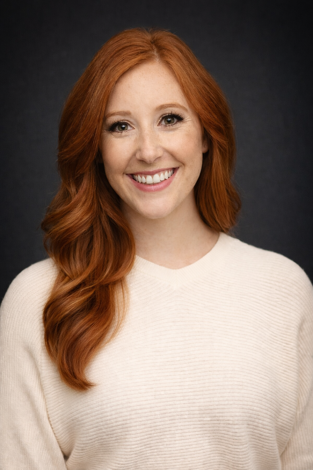 Portrait of a woman with long red hair, blue eyes, and freckles, smiling, wearing a light beige sweater, against a dark background.