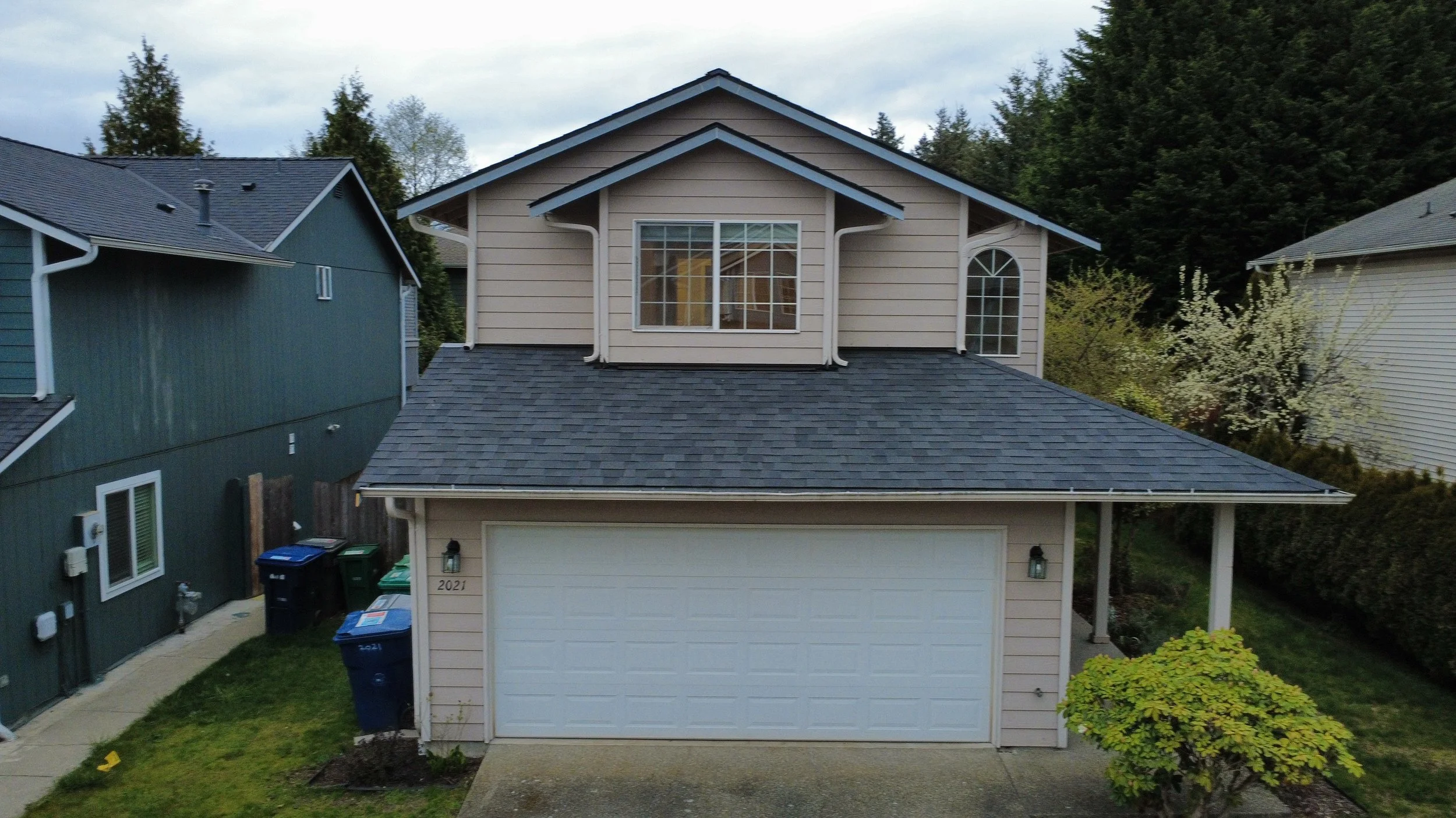 Front view of a two-story house with beige siding, white trim, and a gray shingle roof, surrounded by neighboring houses and trees.