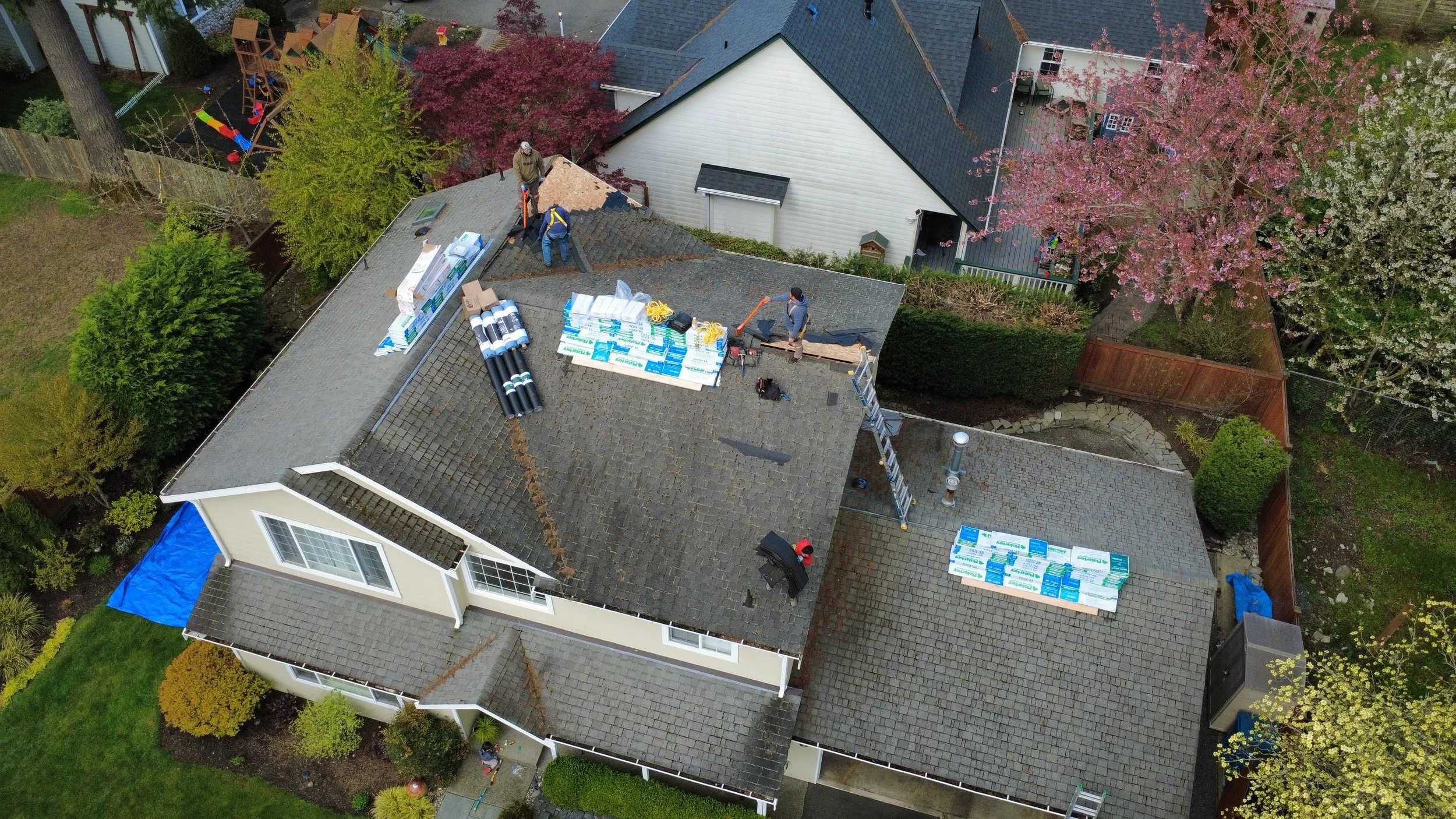 Aerial view of a house with a roofing project in progress, showing workers installing new roofing materials, with supplies and tools on the roof and surrounding yard, neighboring houses visible, and blooming trees.