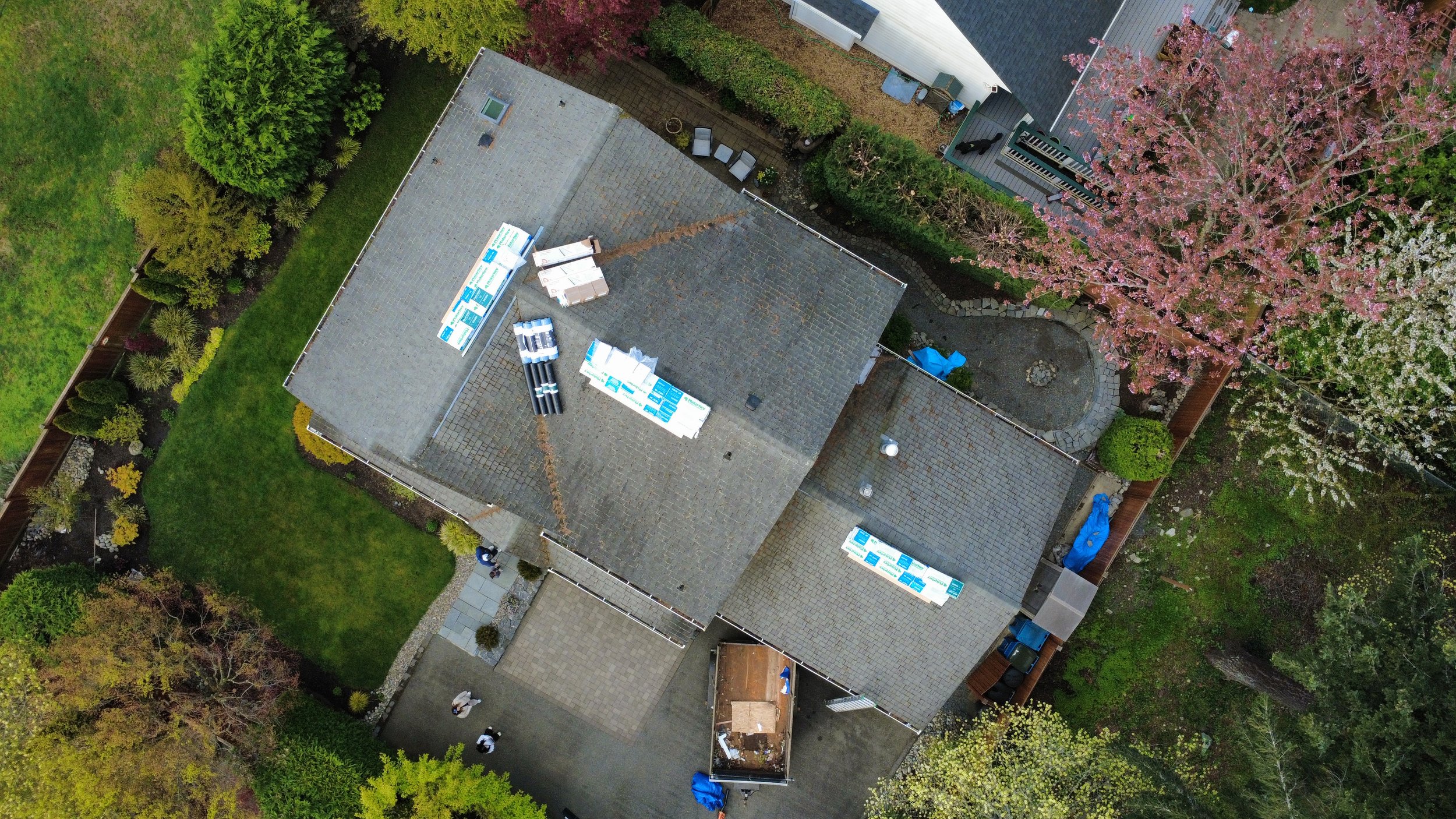 An aerial view of a house with a gray shingle roof, surrounded by green trees and a landscaped yard with flowering trees.