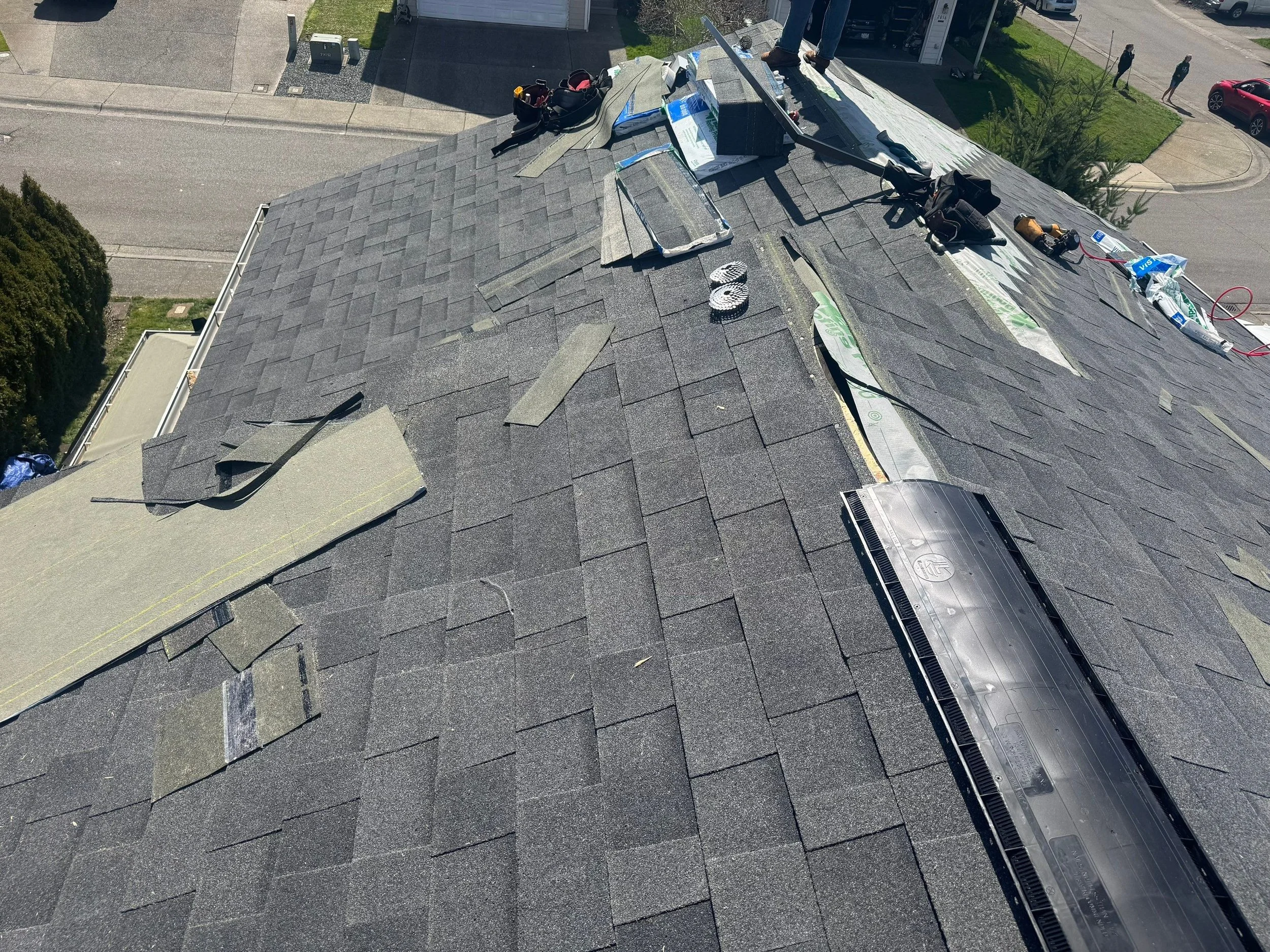 A rooftop undergoing repair, with shingles, tools, and materials scattered across the surface, and a bright sunny day with a few pedestrians and cars visible on the street below.