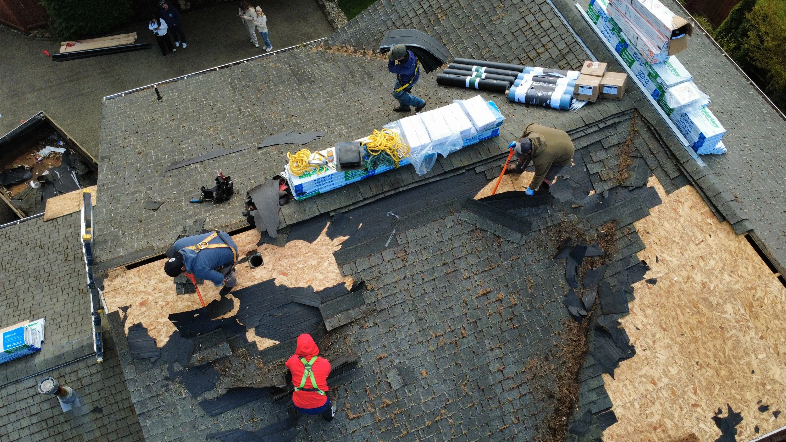 Workers replacing the roof shingles on a house with black asphalt shingles, some shingles removed revealing plywood underneath.