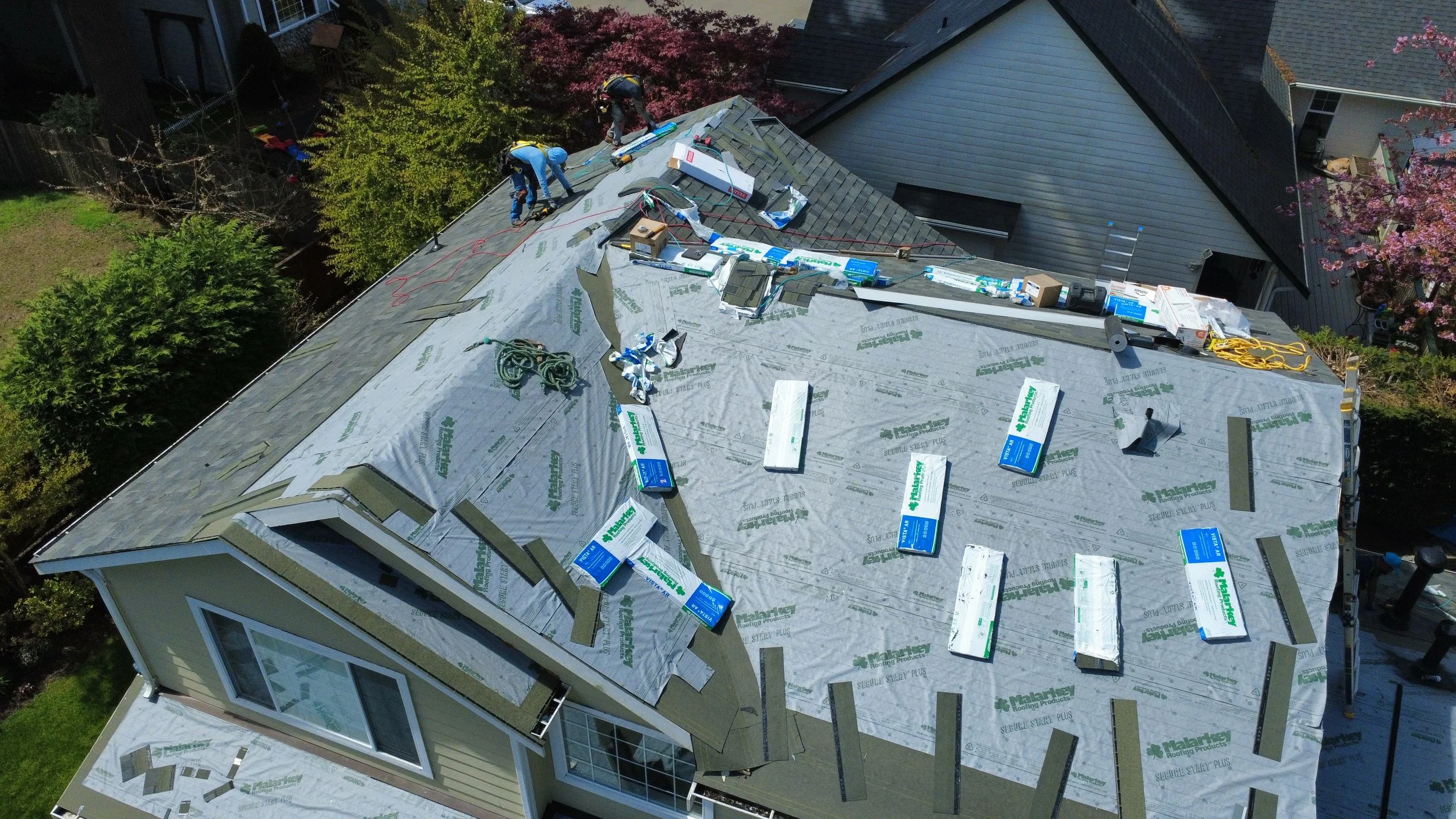 An aerial view of a house's roof under construction with workers installing new roofing materials. The roof is covered with underlayment and some shingles are partially installed, with tools and materials scattered around.