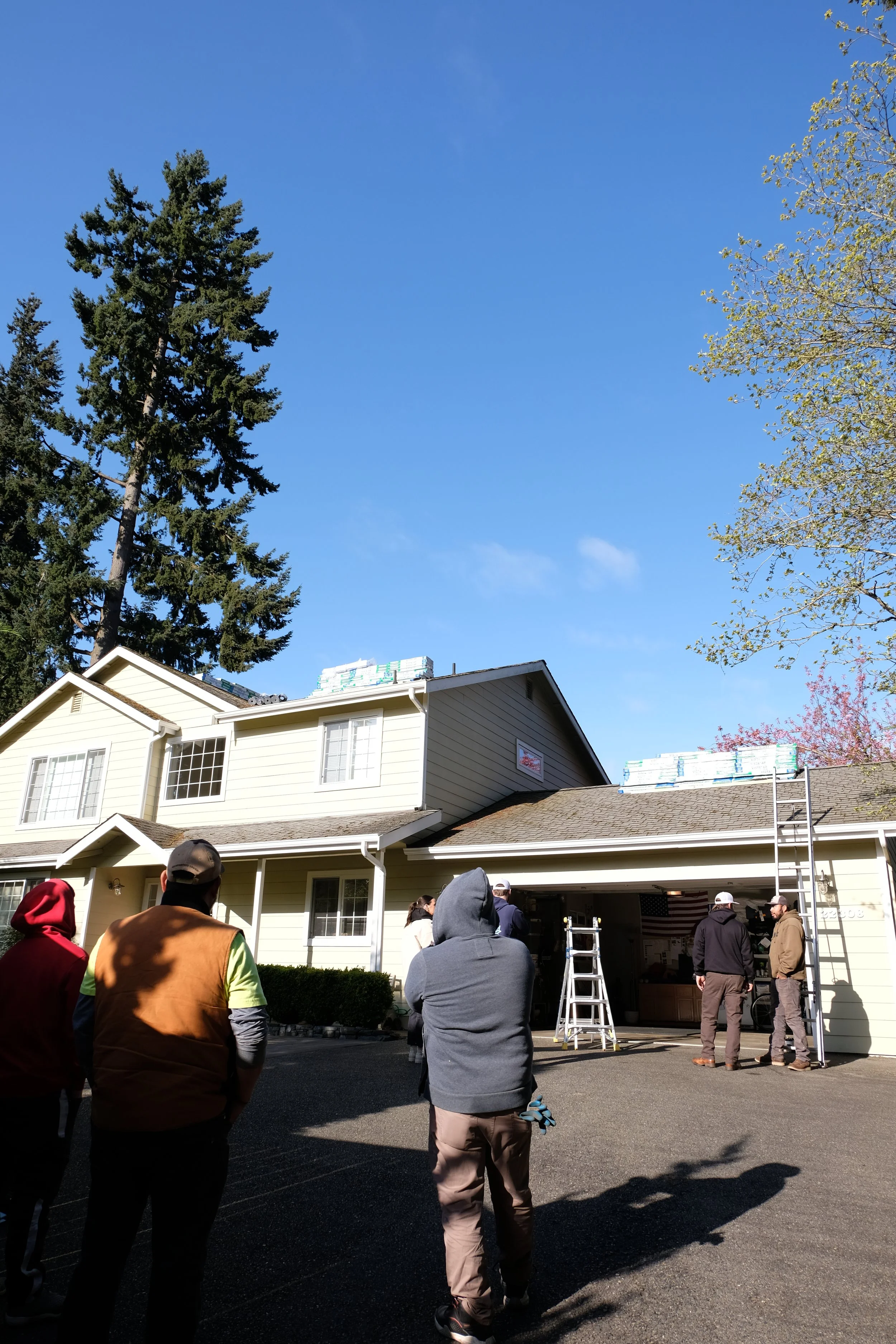A group of people standing outside a two-story house with construction materials on the roof, under a clear blue sky, with tall trees around.