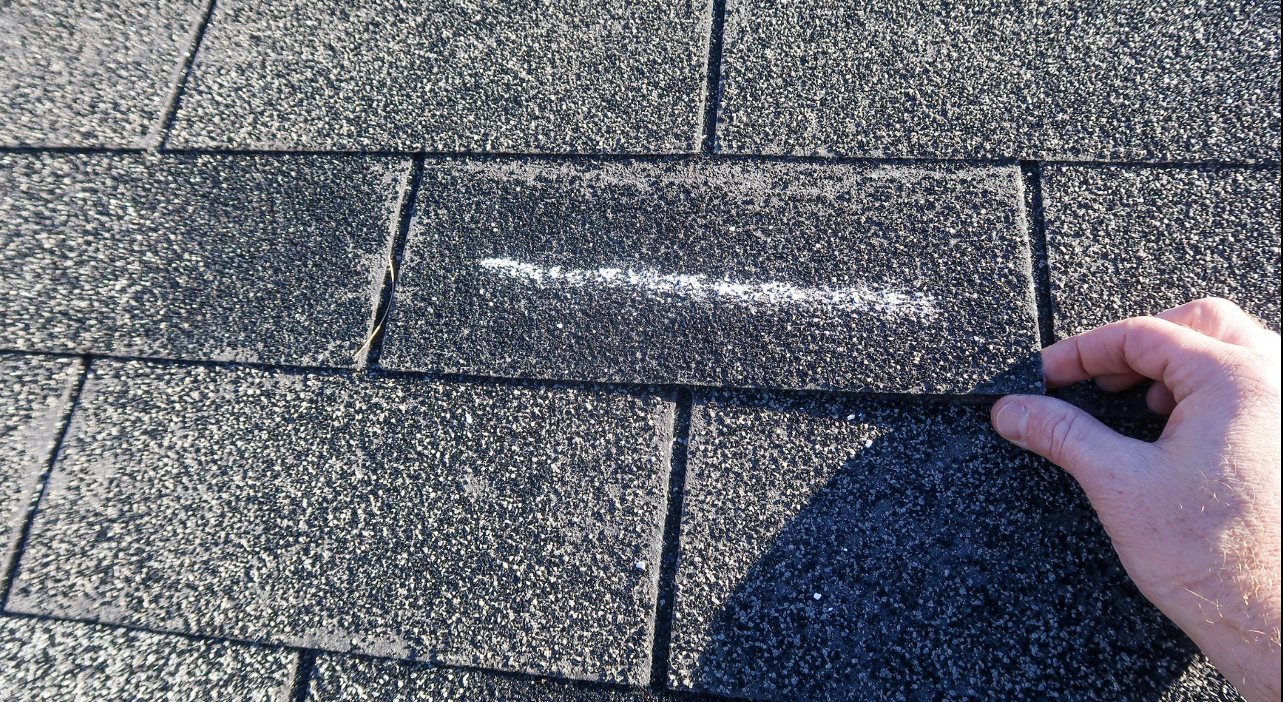 A hand lifting a roofing shingle, revealing a chalk line drawn on the shingles to guide installation.