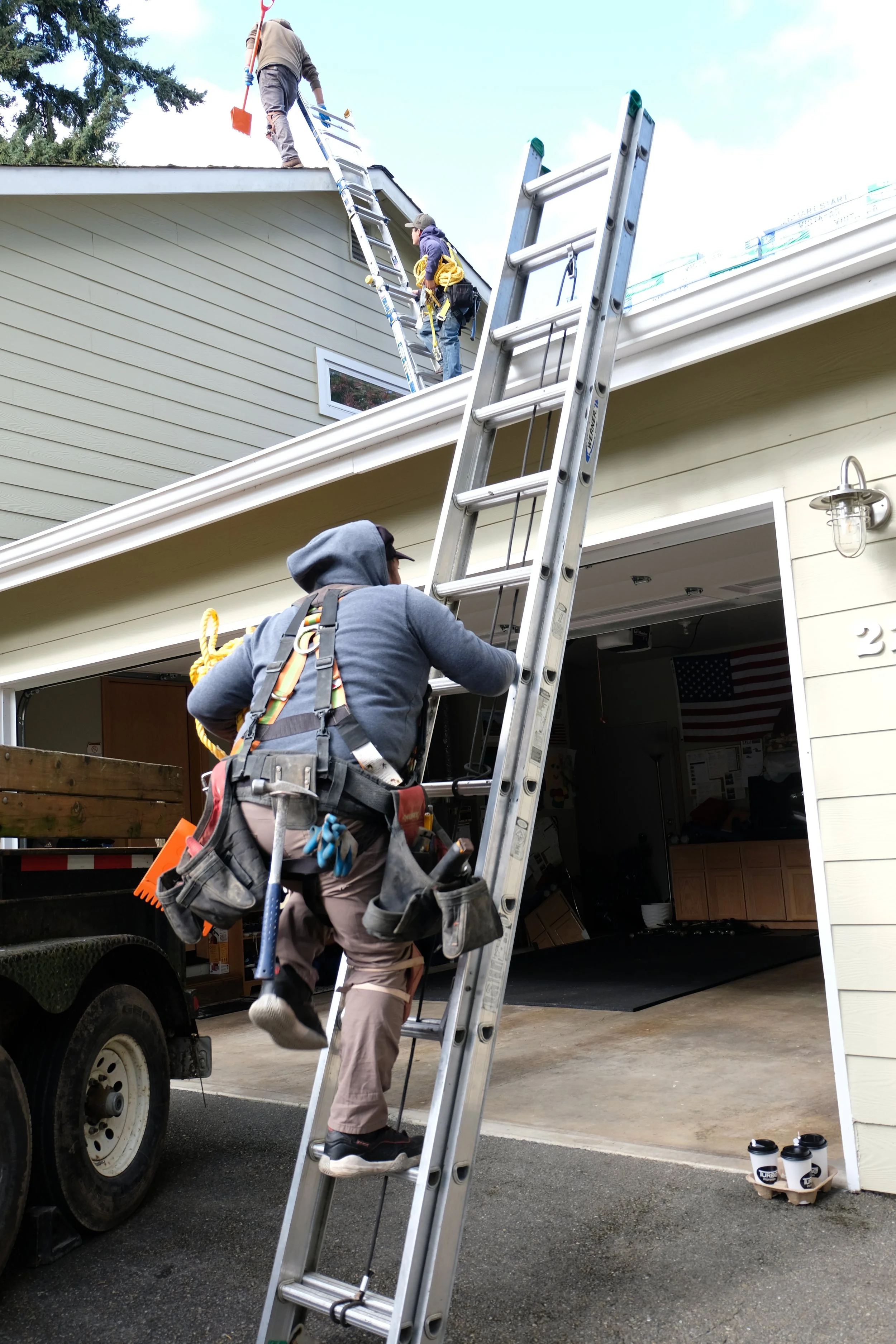Three workers are on a ladder re-roofing a house. One worker is on the roof holding a tool, and two are climbing the ladder. The garage of the house is open inside, showing some items and an American flag hanging. There are coffee cups placed on the driveway.