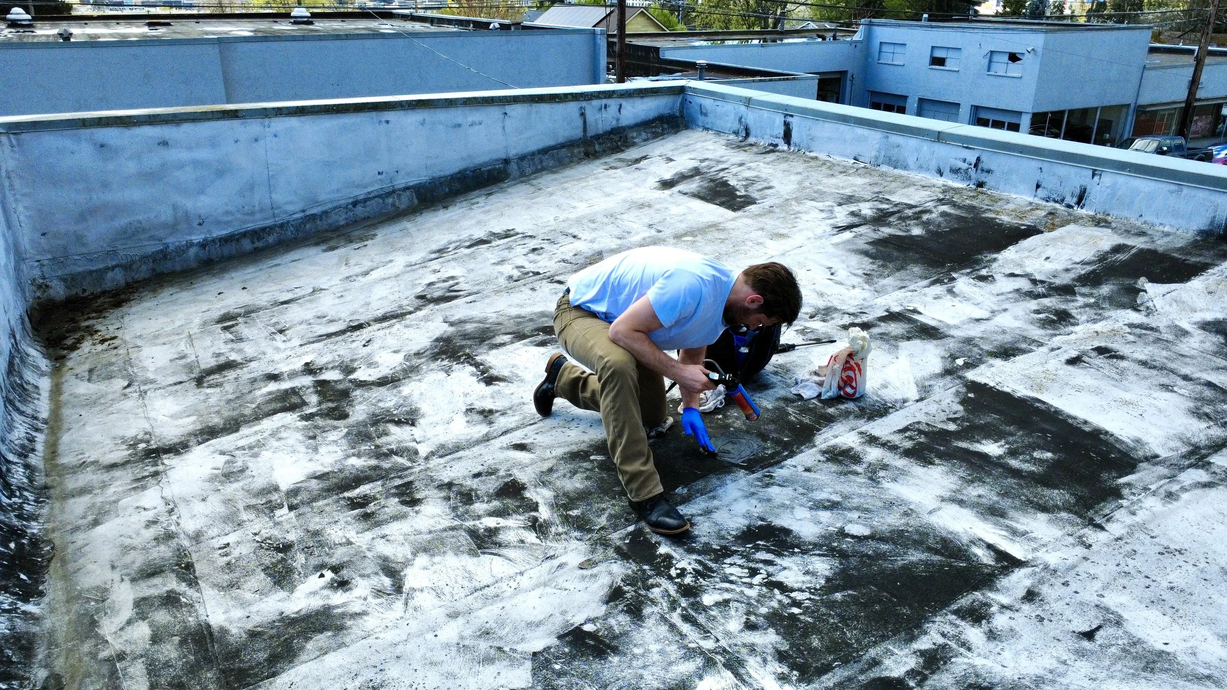 A man in a blue shirt and khaki pants kneels on a rooftop surface covered with black and white tar material, wearing blue gloves, working with tools and a red and white plastic bag nearby. The rooftop has blue walls, and there are buildings and power