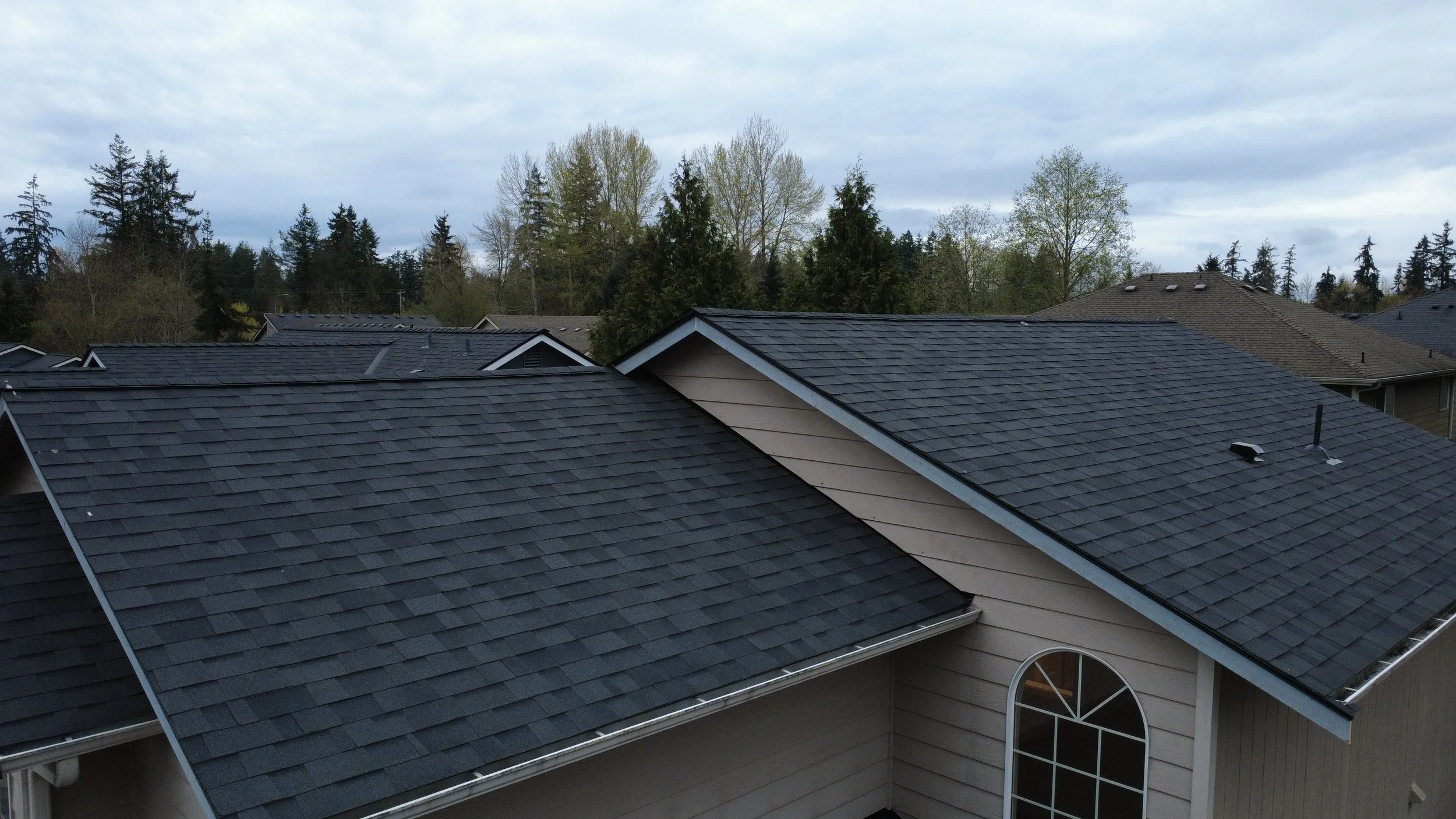 View of multiple houses with dark gray shingle roofs and trees in the background under a partly cloudy sky.
