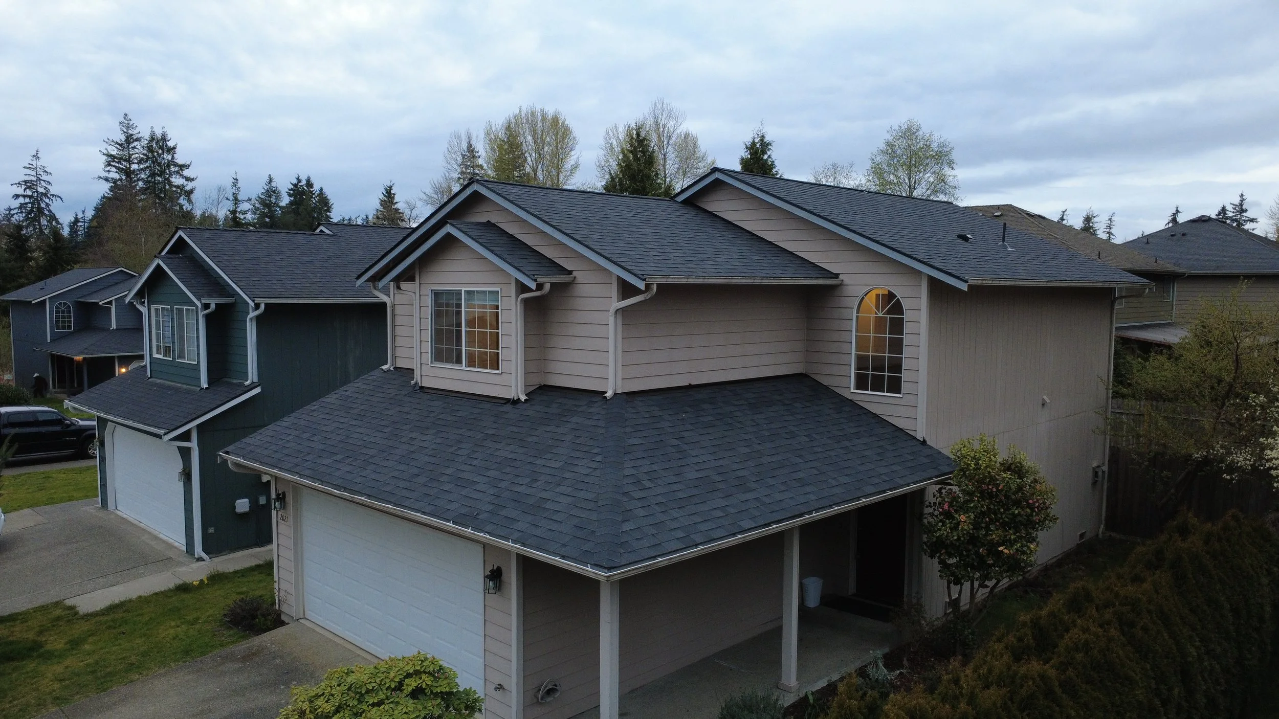 Aerial view of multiple suburban houses with dark gray shingle roofs, some with large arched windows, surrounded by trees and yards.