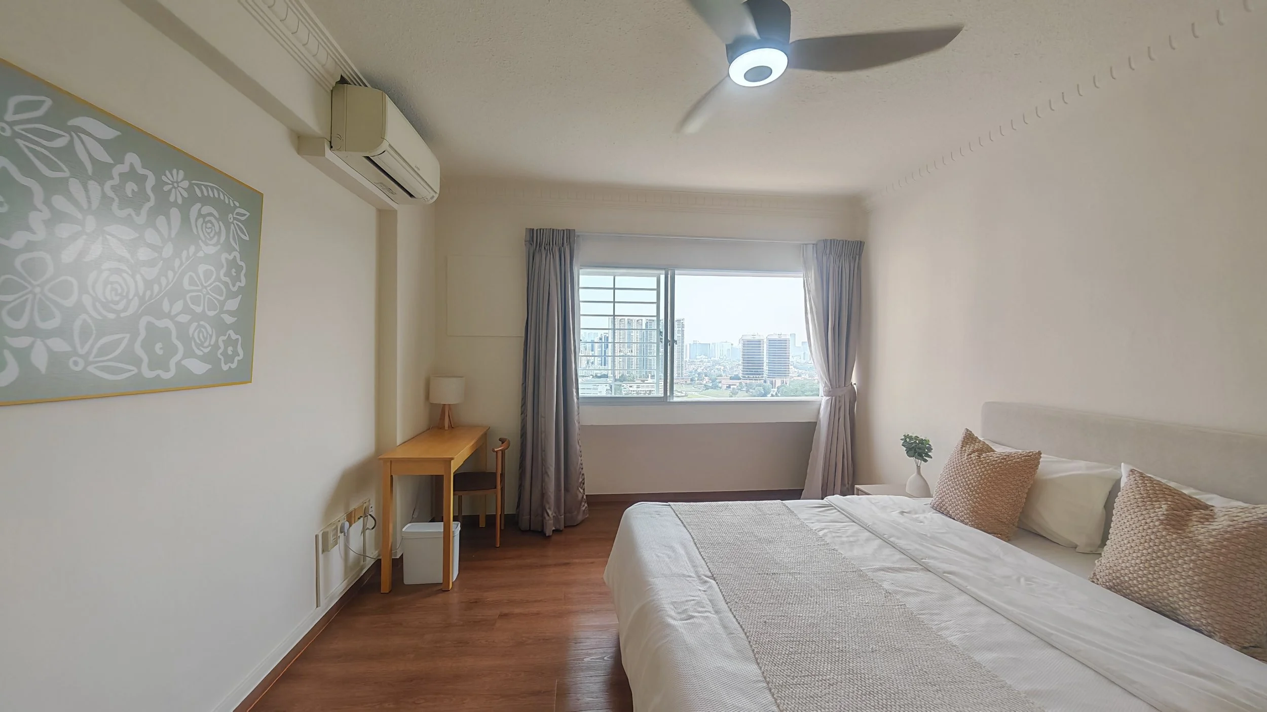 A bedroom with a large bed with white bedding and two beige pillows, a small bedside table with a potted plant, a window with city skyline view, curtains, a wooden desk with a lamp and chair, an air conditioning unit, a ceiling fan, and artwork on the wall.