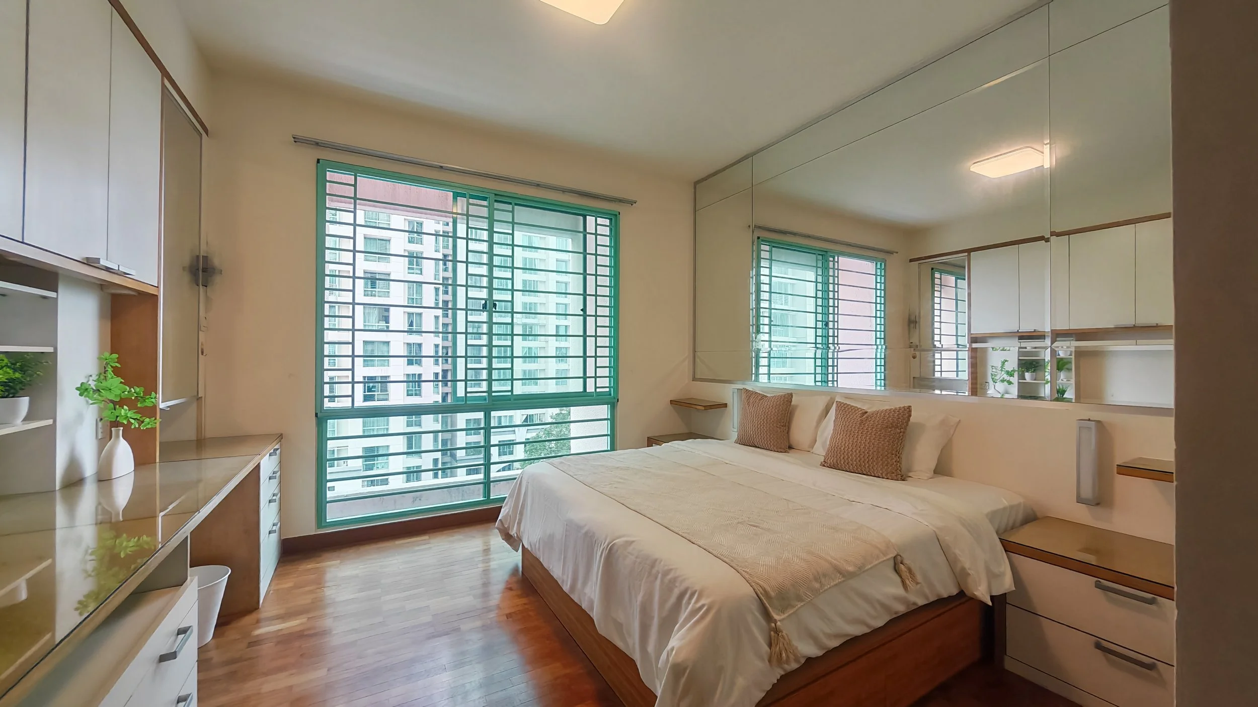 A bedroom with a large bed, pillows, and a blanket, next to windows with green security bars, and a mirror reflecting the room's interior.