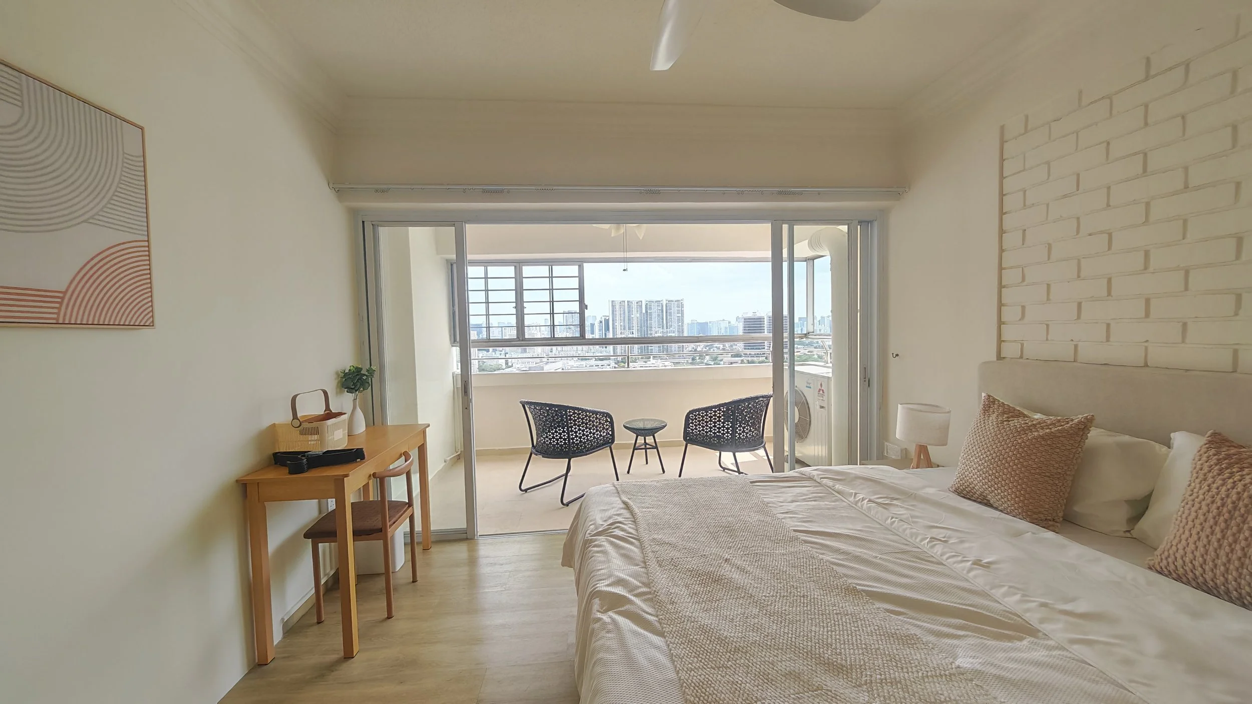 A bedroom with a sliding glass door leading to a balcony, overlooking a cityscape. The room has a bed with beige pillows, a wooden desk with a chair, and a small table with two chairs outside on the balcony.