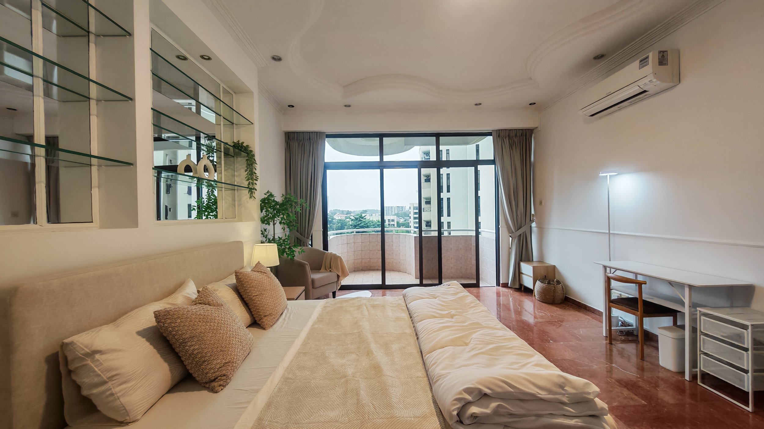 A spacious bedroom with a large bed, beige headboard, and decorative pillows. There is a balcony with sliding glass doors, curtains, and outdoor seating. The room has a white desk, a chair, a floor lamp, and some shelves on the wall. The floor is marble or tile, and the room is well-lit with natural light.